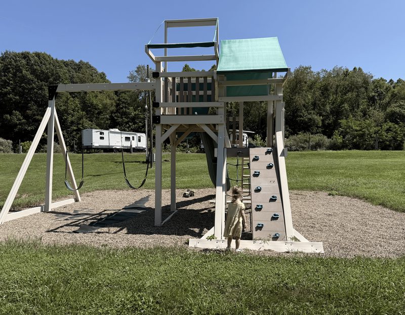 A child at the campground playground with swings, a climbing wall, and a slide.