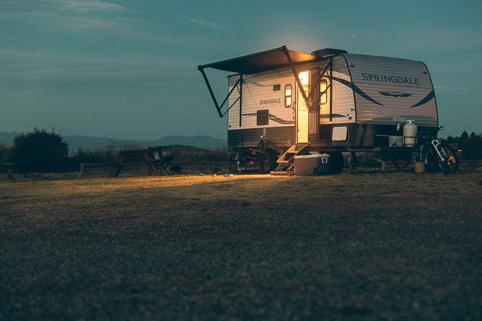 An RV parked at a State Line Campground site at dusk with warm interior lights glowing and hills in the distance.